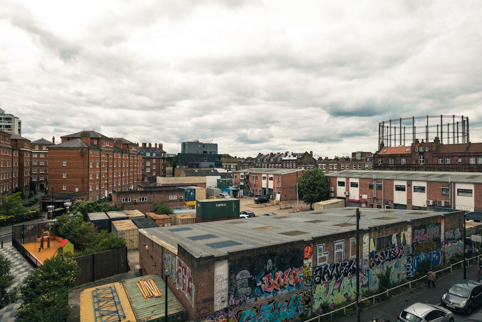A wide urban view showing a mixture of low-rise commercial and residential buildings under a cloudy sky. In the foreground, a flat-roofed brick building features a graffiti-covered wall along its side, with vibrant, colorful spray art. Behind it, there are several small structures with metal and wooden roofs, some surrounded by fencing and greenery, indicating a storage or yard area. To the left, multi-storey brick residential buildings with red brick facades and chimneys are visible, while on the right, a tall circular metal frame structure is situated atop a building. The scene includes parked cars along the street and scattered trees, with a general atmosphere of a densely built-up, urban environment. The image, associated with rubbish removal services by House Clearance Hackney, suggests an area where private waste clearance or alternative waste handling might be relevant, especially for property or commercial clearance purposes.