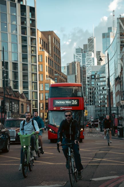 A busy city street scene featuring a group of cyclists riding across a wide road, with three men prominently visible in the foreground. The cyclists are dressed in casual clothing, with some wearing backpacks, and are riding various types of bikes. Behind them, a red double-decker bus displaying the route number '26' and destination 'Hackney Wick' is traveling towards the camera. The background showcases tall, modern office and residential buildings with glass facades and a mix of concrete and brick exteriors, reflecting the urban environment. Overhead, the sky is partly cloudy with patches of blue visible, and the area is well-lit by natural daylight. Surrounding vehicles, including parked cars, are visible along the street edges, and pedestrian walkways are seen at the sides. The scene captures a typical central London intersection, illustrating urban transportation and the mix of private and public mobility options, often associated with alternative waste handling and rubbish removal services in city environments, such as those offered by House Clearance Hackney.