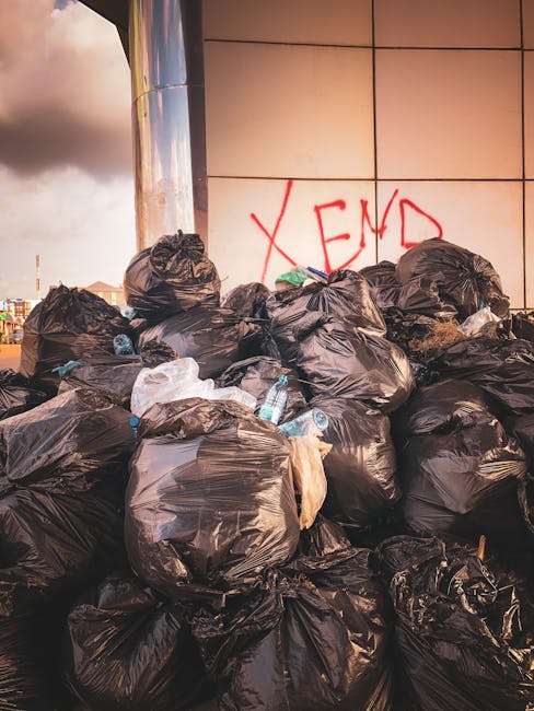 A large pile of black rubbish bags filled with waste is stacked against a tiled exterior wall of a building, with some bags appearing slightly torn or bulging. Several discarded plastic water bottles and miscellaneous rubbish are visible protruding from the bags, indicating general household or domestic waste. The background shows a cloudy sky and an urban environment with faint structural elements, suggesting an outdoor location on a pavement or alleyway. Red graffiti spelling 'XEN' is spray-painted on the wall behind the trash. The scene represents an example of unmanaged refuse accumulation, possibly related to private rubbish collection or alternative waste disposal methods, which some households or commercial premises like House Clearance Hackney might arrange for on-site clearance or independent collection services. The overall atmosphere highlights the challenges of waste management in densely populated areas, emphasizing the importance of reliable rubbish removal to prevent illegal dumping or environmental hazards.