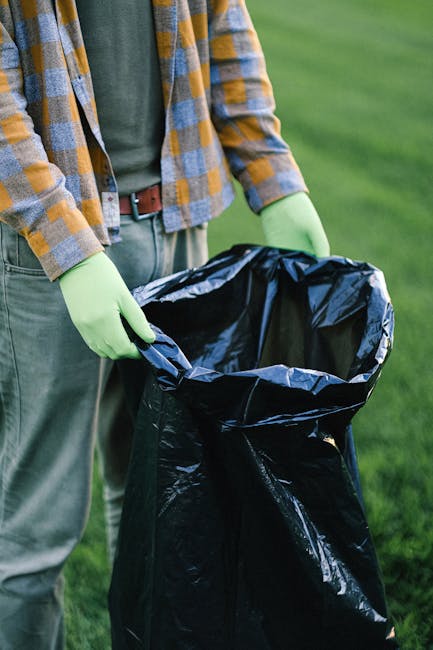 A person wearing a green jacket and yellow plaid shirt is holding open a large black garbage bag with green rubber gloves. The bag, made of shiny plastic material, is positioned in front of a grassy outdoor area with a blurred green background. The individual appears to be preparing the bag for waste collection or rubbish removal, and part of their beige trousers and brown belt are visible. The scene suggests an activity related to private waste handling, such as outdoor rubbish collection or cleanup, consistent with the services offered by House Clearance Hackney, which provides alternative waste collection and on-site clearance solutions.