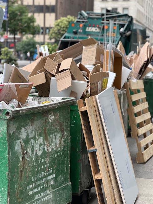 A large green waste bin, positioned on a city street, filled with various discarded cardboard boxes, some flattened and others partially assembled, along with pieces of packaging and packaging materials. The bin shows signs of wear, with scratches and smudges visible on its surface. Surrounding the bin are wooden pallets leaning against it, providing support or for transporting the waste. In the background, a white and green rubbish collection vehicle is partially visible, parked near the curb, with urban buildings and some trees in the distance. The scene suggests an on-site or private waste disposal activity, typical of a rubbish removal or house clearance service, with the waste materials prepared for collection by a service such as House Clearance Hackney. The lighting appears natural, indicating daytime, with a slightly overcast sky contributing to diffuse shadows, emphasizing the textures of the cardboard, pallets, and the waste container, all consistent with a typical urban rubbish clearance scene in Hackney.
