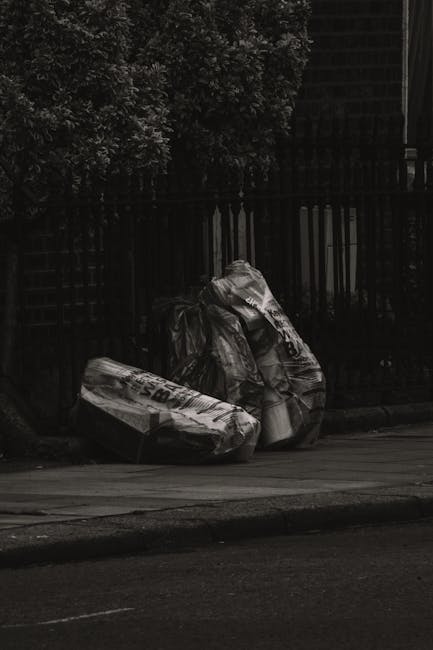 A person wearing a green jacket and yellow plaid shirt is holding open a large black garbage bag with green rubber gloves. The bag, made of shiny plastic material, is positioned in front of a grassy outdoor area with a blurred green background. The individual appears to be preparing the bag for waste collection or rubbish removal, and part of their beige trousers and brown belt are visible. The scene suggests an activity related to private waste handling, such as outdoor rubbish collection or cleanup, consistent with the services offered by House Clearance Hackney, which provides alternative waste collection and on-site clearance solutions.