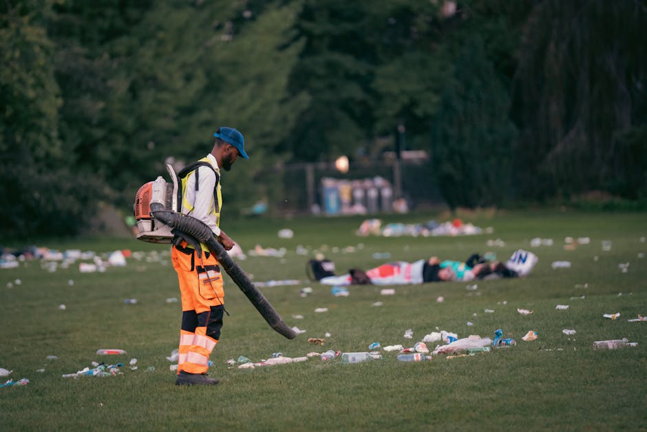 A worker wearing a blue cap, reflective orange and black trousers, and a safety vest is using a leaf blower or vacuum to clear a grassy area covered with scattered litter, including plastic bottles, cups, and paper debris. The scene takes place outdoors, with dense green trees and bushes in the background, and some trash bags and waste collection points visible further back. The worker is positioned to the left of the image, with the scattered rubbish spread across the foreground and middle ground, indicating a recent event or gathering requiring cleanup. This scene exemplifies private waste handling and rubbish removal efforts, supported by professional waste management services such as House Clearance Hackney, which manage on-site clearance tasks on behalf of clients. The natural lighting suggests early evening or late afternoon, highlighting the importance of thorough and efficient rubbish removal in maintaining clean outdoor spaces after large public or private events.
