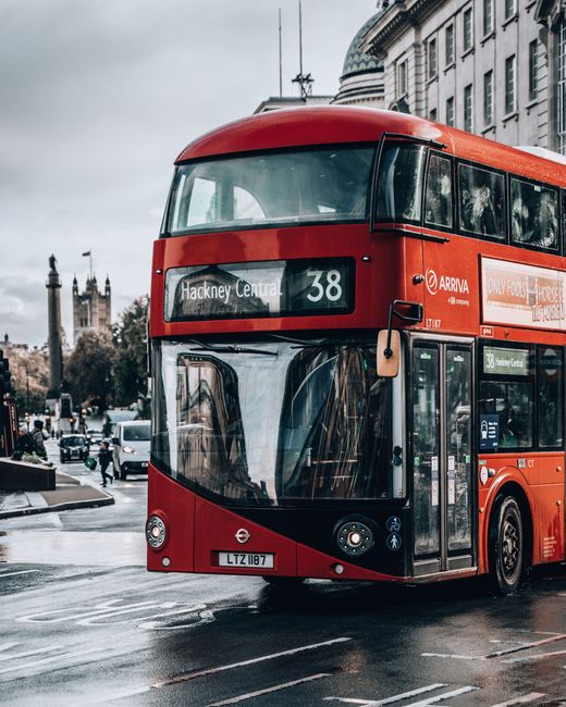 A red double-decker bus operating on a London city street, with the route number 38 and destination Hackney Central displayed on the front. The bus is parked near the pavement, reflecting the surrounding urban scenery in its large front windows. In the background, historic buildings with ornate architectural details and clock towers are visible, along with trees and other vehicles on the wet road. The overcast sky and damp pavement indicate recent rain, contributing to a subdued, professional city atmosphere. The bus's exterior has a smooth, glossy finish, and the vehicle features typical London transport branding and signage. This scene exemplifies typical urban transport infrastructure, relevant in contexts of local and private transport or alternative waste handling arrangements in city environments, as discussed in rubbish removal and clearance services managed by companies like House Clearance Hackney.