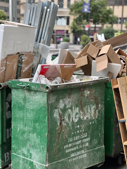 A large outdoor area features numerous stacks of cardboard fruit and vegetable boxes, primarily marked with the 'fresh fruits' label, arranged in various heights and occupying a significant portion of the space. To the left, there are two green plastic waste bins with closed lids, positioned on a paved surface. Several black plastic crates and a red shopping trolley filled with black and blue trash bags are visible in the foreground, alongside a smaller red plastic container. On the right, there are several metal wire cages filled with flattened cardboard boxes and assorted packaging materials, with some boxes showing labels and barcodes. The background reveals a rural setting with a pond, lush green trees, and a few houses with sloped roofs under a clear sky, suggesting a suburban or semi-rural environment. The scene is illuminated by natural daylight, emphasizing the textures and colors of the various waste materials, which appear to be awaiting collection or disposal. The image is associated with private waste handling or on-site clearance, consistent with services provided by House Clearance Hackney.