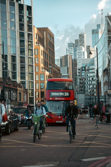 A busy city street scene featuring a red double-decker bus with a digital display indicating route 26 to Hackney Wick, positioned centrally amidst modern and traditional high-rise buildings with glass and brick facades. The street has multiple pedestrians, some walking and others cycling on designated bike lanes, including a man in dark clothing riding a bicycle in the foreground. Several cars and motorcycles are visible on the left side of the image, parked along the curb. The weather appears clear, with a partly cloudy sky overhead, providing natural lighting that highlights the urban environment. The surrounding environment emphasizes an active cityscape with a mix of commercial and residential structures, typical of central London areas. The image subtly connects to urban waste management themes, as it captures a snapshot of city life where efficient rubbish removal and planning are essential for maintaining a clean and functional urban setting, aligning with the services offered by House Clearance Hackney.