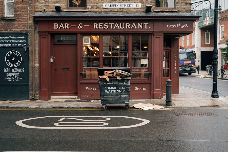 A black wheeled commercial waste bin positioned on the sidewalk directly in front of a red-painted storefront, which is a bar and restaurant with large glass windows and wooden framing. The bin is filled with flattened cardboard boxes and other waste materials, some overflowing onto the ground. The signage on the bin indicates it is designated for 'Commercial Waste Only.' The facade of the establishment features signage advertising wines, spirits, and cocktails, with a small blackboard on the left advertising a self-service buffet open weekdays from 11am to 3pm. A black bollard is situated to the right of the bin, and a black lamppost is also visible nearby. In the background, a delivery truck and bicycles are parked at the street corner, with residential buildings and street signs visible further back. The scene captures a typical urban setting where private waste collection and rubbish disposal are necessary for commercial premises, supporting services like independent rubbish removal consistent with local regulations and waste management practices.