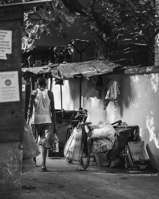 A young man walking along a narrow alleyway captures an urban scene in black and white. He is dressed in a sleeveless shirt and shorts, holding a plastic bottle in one hand while carrying a bottle of water in the other. To his right, there's a bicycle loaded with various types of rubbish, including large bags filled with debris and some loose items, which appear to be waste materials awaiting collection. Behind the bicycle, a makeshift shelter constructed from wooden poles and tarpaulin offers partial cover, with additional trash and discarded objects piled underneath or nearby. The alley is bordered by a high wall on one side, with a small section of foliage visible at the top, creating dappled shade. The environment appears cluttered and congested, characteristic of informal waste accumulation points that may require private rubbish removal services like those offered by House Clearance Hackney. Sparse natural light filters through the trees, casting uneven shadows over the scene, emphasizing the documenting nature of this independent waste collection effort in an urban setting.