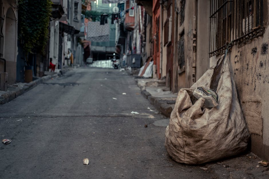 A large, weathered, off-white fabric sack filled with mixed waste materials, including a clear plastic container, resting on the uneven asphalt pavement of a narrow, urban alleyway. The sack appears dirty and slightly sagging, with its top folded over and the plastic container partially visible at the opening. The surrounding environment features closely packed residential buildings with graffiti and black metal window grilles on the right side, and a dark, cluttered street extending into the distance with hanging laundry, makeshift repairs, and scattered debris, indicating an area where independent rubbish collection or site clearance services may be required. Natural lighting suggests overcast weather, casting soft, diffuse light over the scene, emphasizing the gritty and neglected atmosphere typical of urban rubbish accumulation needing professional waste management from companies like House Clearance Hackney.