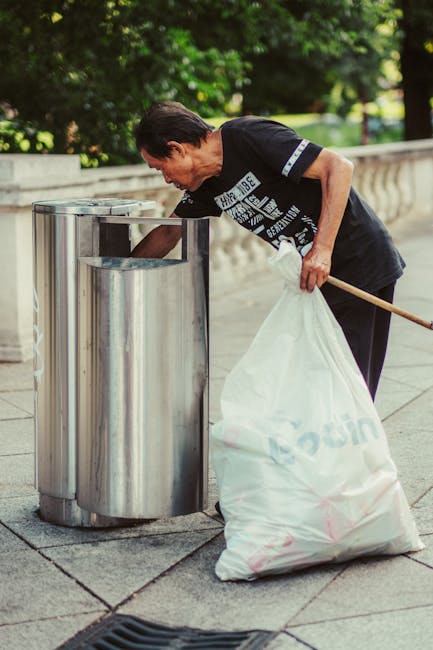 A middle-aged man with dark hair, dressed in a black T-shirt and dark trousers, is seen disposing of waste into a tall, shiny stainless steel public litter bin on a paved pathway. The man leans forward slightly, holding a long wooden stick or pole in his right hand to assist in emptying a large white plastic bag filled with rubbish. The bag appears to contain various waste items, and the man's focus is directed toward the open top of the bin as he manages debris into it. The setting is outdoors, with a background of lush green trees and a stone balustrade, indicating a park or garden area. The scene is illuminated by natural daylight, creating a bright and clear atmosphere suitable for public waste disposal or maintenance activities. This image subtly illustrates an acceptable alternative to on-site rubbish collection, demonstrating waste handling and collection that could be associated with private or independent rubbish removal services, such as those offered by House Clearance Hackney.
