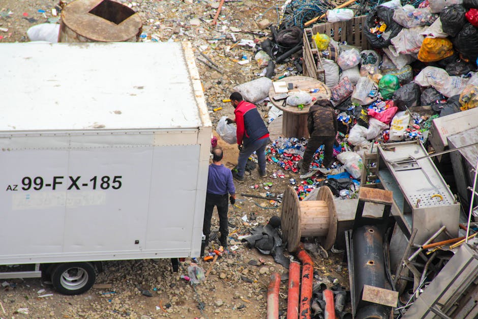 An aerial view of a significant amount of mixed rubbish and waste materials scattered on the ground in an outdoor area. Visible are various black, white, and patterned plastic bags filled with waste, alongside loose trash including paper, plastic bottles, and packaging. A large white delivery truck marked with the code '99-FX-185' is parked on the left side, partially blocking the view of the waste. Three individuals are actively engaged in sorting or clearing the debris; one person in a red and black jacket is bending down, another in a dark outfit is leaning over, and the third, with a gray top, is closer to the truck. The surrounding environment features an uneven, gravelly surface with scattered smaller rubbish, large wooden spools, and broken equipment or piping, indicating a site of informal waste collection or cleanup. The scene exhibits natural daylight, with a focus on manual rubbish handling that could be part of private waste disposal or independent cleanup services in Hackney, as provided by companies like House Clearance Hackney.