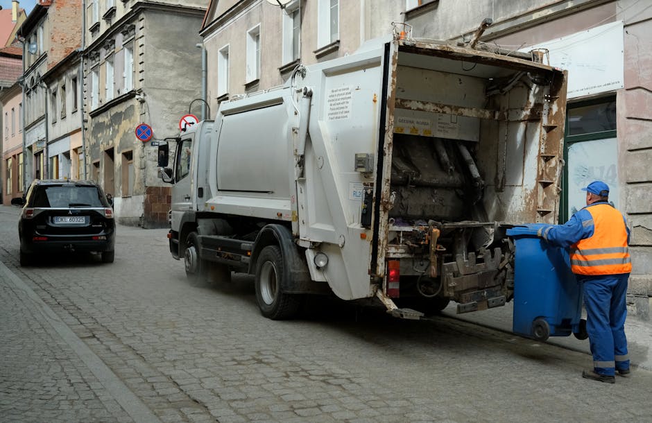 A waste collection truck with an open rear compartment positioned on a narrow urban street, adjacent to weathered, multi-story buildings with mixed architectural details. The truck is white with some visible rust and grime, indicating frequent use, and features a large hydraulic lift mechanism at the back for loading waste. A worker wearing a blue uniform, orange high-visibility vest, and a blue cap is seen placing a blue wheeled bin into the truck’s rear, preparing for rubbish removal or an onsite clearance. The street’s cobblestone pavement is partially visible, with parked cars along the roadside, including a black vehicle parked behind the waste truck. The environment suggests an individual waste collection or private rubbish removal process, aligned with services like those offered by House Clearance Hackney, supporting alternative waste handling outside of municipal collection systems. The scene is well-lit, typical of daytime street operations, emphasizing the practical nature of rubbish removal services in urban environments.